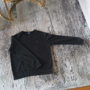 Svart tröja från Polo Ralph Lauren - Svart långärmad tröja från Polo Ralph Lauren med klassisk rund hals och röd broderad logga på bröstet. Tröjan är i mjuk bomull och har en clean, stilren look som funkar till allt. Perfekt basic i garderoben för dig som gillar enkel och snygg stil.