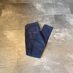 Mörkblå jeans från Nudie Jeans - Säljer ett par mörkblåa jeans från Nudie Jeans i klassisk femficksmodell. Jeansen har smal passform, normal midja och är tillverkade i stretchig denim för extra komfort. Perfekta för dig som gillar en clean och stilren look.