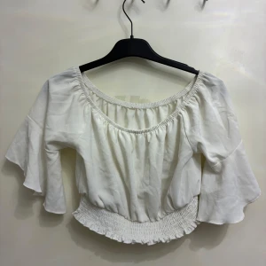 Vit croppad blus med volangärm - Superfin vit blus med vida volangärmar och smockad midja. Offshoulder-modell med elastisk halsringning som ger en snygg och avslappnad look. 