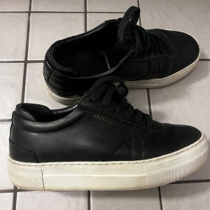 Svarta sneakers från Axel Arigato - Stilrena svarta sneakers från Axel Arigato med vit platt sula och diskret guldfärgad logga på sidan. Skorna är i skinn och har klassisk snörning. Perfekta för dig som gillar minimalistisk och clean stil.