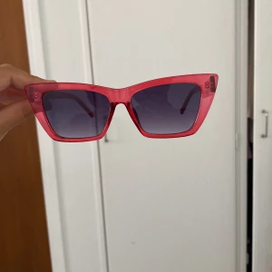 Rosa cat eye solglasögon från Glitter - Snygga solglasögon i cat eye-modell från Glitter. Ramen är transparent rosa och glasen är mörka med en lätt toning. Perfekta för att ge din look en cool och trendig vibe. Lätta och bekväma att bära hela dagen.