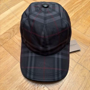 Rutig keps - Snygg keps från  London i mörkgrått och svart med röd och grå rutig design. Klassisk böjd skärm och justerbar rem baktill i läder med präglad logga. Tillverkad i slitstark polyester, perfekt för en trendig streetstyle.