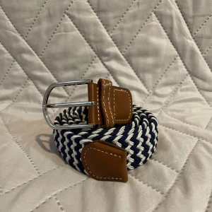 Randigt flätat bälte med läderdetaljer - Snyggt flätat bälte i marinblått och vitt med bruna läderdetaljer och silverspänne. Perfekt accessoar för att lyfta din outfit med en sportig och stilren touch. Bältet har tydliga kontrastsömmar på lädret och är flexibelt i materialet. Den är 100cm lång.