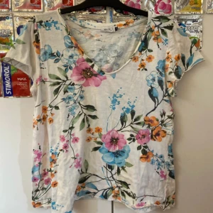 Blommig t-shirt från Mix by Heart - Vit t-shirt från Mix by Heart med färgglatt blommönster i blått, rosa och orange. T-shirten har korta ärmar och v-ringning. Materialet känns mjukt och luftigt, perfekt för en chill look. Snygg passform som är lätt att matcha med jeans eller shorts.