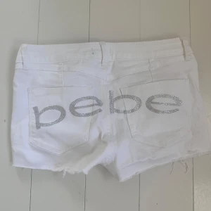 Vita jeansshorts med strass från Bebe - Snygga vita jeansshorts från Bebe med råa kanter och låg midja. Baksidan har stora bakfickor med glittrande strassbokstäver som stavar 'bebe'. Klassisk femficksmodell med dragkedja och knapp 💋