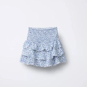 Blommig volangkjol i blått och vitt - Supersöt kort kjol med inbyggda shorts och blått blommönster på vit botten. Kjolen har flera volanger och smock i midjan för en bekväm passform. Perfekt för varma dagar när du vill känna dig extra söt och somrig. Storlek 128 barn för att få kortare volanger men passar som XS 🩵🤍💙