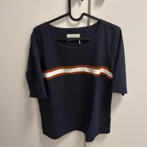 Mörkblå topp med rand från Freequent - Mörkblå topp från Freequent med kort ärm och rund hals. Framtill finns en horisontell rand i vitt och brunt som ger en sportig vibe. Skön och lätt i materialet, perfekt till jeans eller kjol för en chill look.