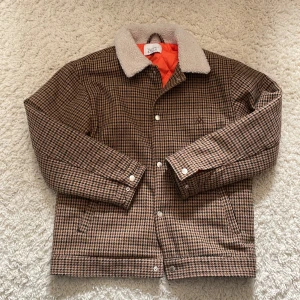Valient jacka - Snygg rutig teddyjacka från Valley Studio med brun och svart färg, vit teddykrage och orange innerfoder. Jackan har knappar framtill och klassisk rak passform. Perfekt för dig som vill ha en retrovibe och värme i höst och vinter.