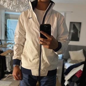 Vit vindjacka från Etirel med blå detaljer - Snygg vit vindjacka från Etirel med marinblå muddar och krage. Jackan har dragkedja framtill, två sidofickor och stilren logga på bröstet. Perfekt för dig som gillar en clean och sportig look. Passar till jeans eller shorts för en chill stil.