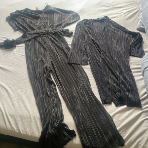 Svart plisserad set - Säljer en svart plisserad byxdress med omlott-topp och vida ben. Materialet är glansigt och mjukt, vilket ger en lyxig känsla. Byxdressen har knytband i midjan och ett snyggt fall. Perfekt för dig som gillar trendiga och bekväma plagg.