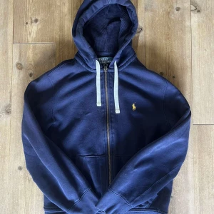 Marinblå hoodie från Polo Ralph Lauren - Snygg marinblå hoodie från Polo Ralph Lauren med dragkedja och vit snörning i huvan. Klassisk gul broderad logga på bröstet och känguruficka framtill. Mjuk bomullskvalitet och långärmad model. Lite använd men ser fortfarande bra ut!