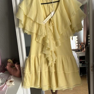 Gul volangklänning från NA-KD - Supersöt ljusgul kortklänning från NA-KD i chiffong med flera lager volanger och knytning fram. Klänningen har en flowy passform och är perfekt för sommarens alla tillfällen. Tillverkad av återvunnet material. Köptes för 599kr.