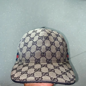 Gucci keps med GG-mönster, M - Snygg keps från Gucci i beige och marinblått med klassiskt GG-monogram över hela kepsen. Har en röd och marinblå rand på sidan och justerbar baksida. Tillverkad i bomullsmix med insida i marinblått tyg. Perfekt för dig som vill sticka ut med lyxig streetstyle. Priset kan diskuteras vid snabb affär 