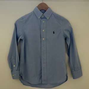 Blå skjorta från Ralph Lauren, Barn 10-11 år - Klassisk ljusblå skjorta från Ralph Lauren i slim fit-modell. Skjortan har knappar framtill, button down-krage och det ikoniska broderade logotypen på bröstet. Tillverkad i mjuk bomull och har långa ärmar med manschettknappar. 