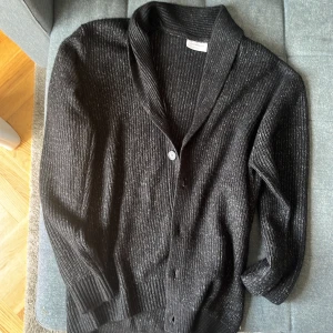 Svart ribbad kofta från Lindbergh - Svart ribbad kofta från Lindbergh med avslappnad passform. Koftan har knappar framtill, bred krage och långa ärmar. Perfekt för lager-på-lager och enkel att styla med olika outfits. Nypris 699 storlek L