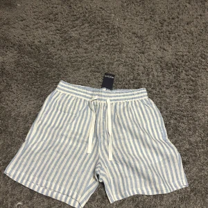 Randiga shorts från Notre Succès - Ljusblå och vita randiga shorts från Notre Succès med elastisk midja och snörning framtill. Tillverkade i ett lätt och luftigt linnematerial, perfekta för varma dagar. Klassisk passform med fickor i sidorna och en bakficka för chill sommarstil.