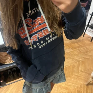 Mörkblå Superdry hoodie med tryck - Säljer en mörkblå hoodie från Superdry med stort broderat och tryckt mönster i rött, vitt och orange på bröstet. Tröjan har huva med vita snören och en klassisk känguruficka framtill. Perfekt för dig som gillar streetstyle.