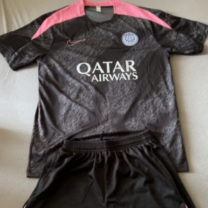 PSG fotbolls set svart/rosa Nike - Snygg PSG fotbolls set från Nike i svart med rosa detaljer på axlarna och klubbmärke på bröstet. Tröjan har korta ärmar och Qatar Airways-tryck på framsidan. Materialet är lätt och andas, perfekt för träning eller match. 