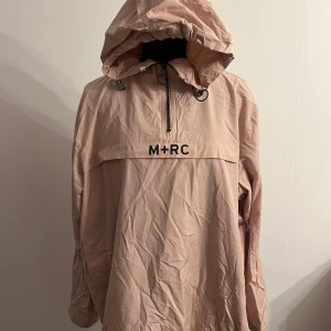 Ljusrosa vindjacka med huva från M+RC NOIR - Säljer en ljusrosa vindjacka från M+RC NOIR med huva och halv dragkedja framtill. Jackan har en avslappnad passform och är tillverkad i ett lätt syntetmaterial. Perfekt för blåsiga dagar och enkel att slänga på sig över en hoodie.
