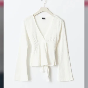 Vit omlottopp med knytband - Säljer en vit omlottopp med v-ringning och knytband i midjan. Toppen har långa ärmar och är tillverkad i ett lätt, strukturerat material. Perfekt för en stilren och fräsch look.