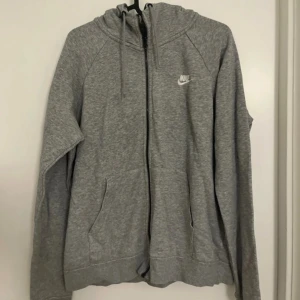 Grå zip-hoodie från Nike - Grå hoodie från Nike med dragkedja och huva. Klassisk design med Nike-logga på bröstet, två fickor framtill och justerbar dragsko i huvan. Perfekt för en avslappnad stil.
