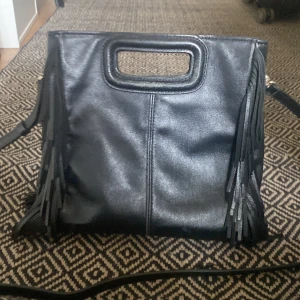 Svart axelväska med fransar från Maje - Snygg svart axelväska i skinn från Maje med fransar på sidorna och ett stilrent handtag upptill. Väskan har en rektangulär form och avtagbar axelrem. Perfekt för dig som vill ha en cool accessoar med lite edge.☀️