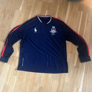 Blå långärmad USA fotbollströja från Ralph Lauren - Mörkblå långärmad fotbollströja med röda ränder längs ärmarna. USA-märke och nummer 2 på ryggen, samt Ralph Laurens logga på bröstet. Vit och blå krage med sportig känsla. Perfekt för dig som gillar klassisk amerikansk stil.