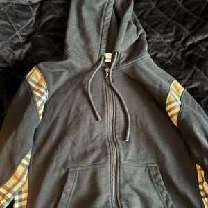 Svart burberry zip - Svart hoodie från Burberry med dragkedja och klassiska rutiga paneler längs ärmarna. Huva med snörning och två fickor framtill. Perfekt för en stilren och avslappnad look.
