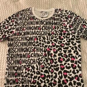 Vit t-shirt med leopardmönster och MOSCHINO-tryck - Säljer en vit t-shirt från Moschino Couture med svart och rosa leopardmönster och MOSCHINO-tryck över hela plagget. T-shirten har rund halsringning och korta ärmar. Perfekt statement-plagg för dig som gillar att sticka ut.