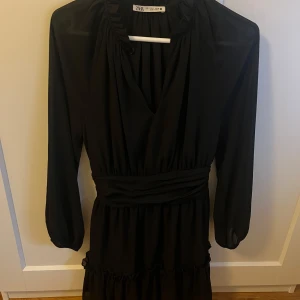Svart långärmad klänning från Zara - Säljer en svart långärmad klänning från Zara med v-ringad omlott framtill och markerad midja. Klänningen har lätt puffade ärmar och volangdetalj nedtill. 