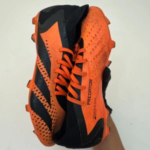 Adidas Predator fotbollsskor orange och svarta - Säljer ett par Adidas Predator fotbollsskor i orange och svart med snörning och mönstrade detaljer. Skorna har en låg profil och är designade för spel på gräs. Perfekta för dig som vill sticka ut på planen!