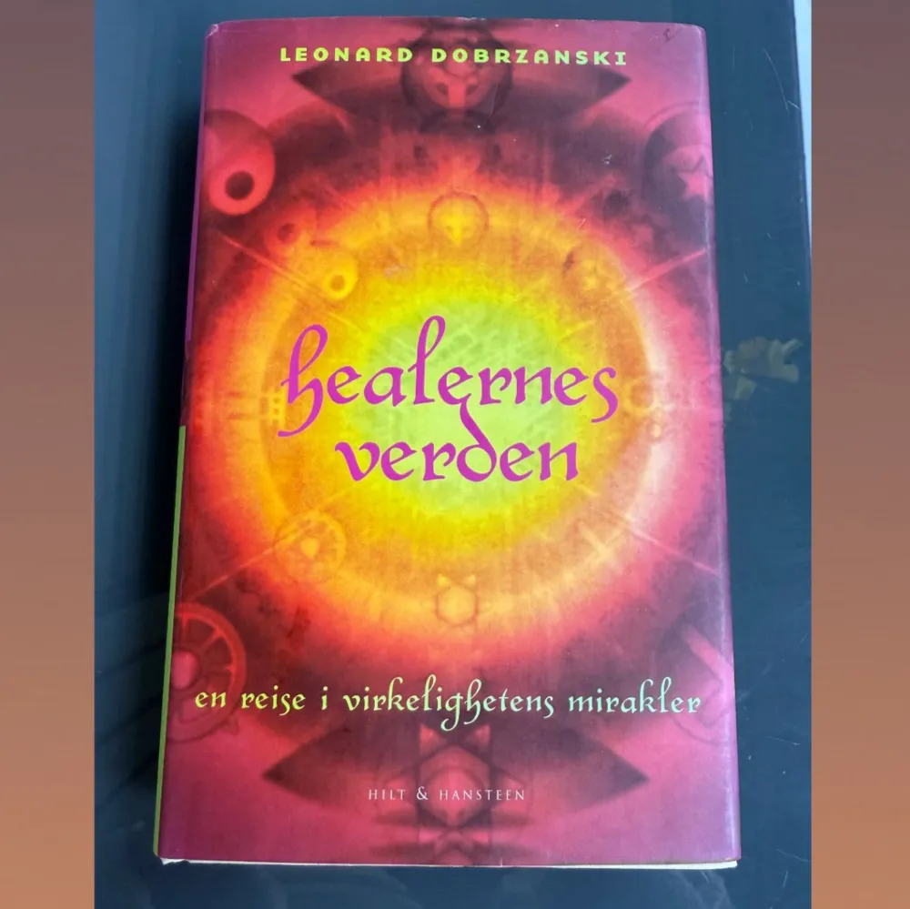 Bok med röd och orange omslag, titeln 'Healernes verden' i lila text. Undertitel: 'en reise i virkelighetens mirakler'. Författare: Leonard Dobrzanski. Boken utforskar healing och alternativa behandlingsformer. Utgiven av Hilt & Hansteen. Bra skriven bok som är skriven på norska. Böcker.