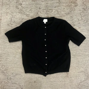 Svart kortärmad kofta från H&M - Säljer en svart kofta från H&M med korta ärmar och knappar framtill. Enkel och stilren modell som passar perfekt till många olika outfits. Mjuk och bekväm, perfekt för lager-på-lager.