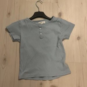 Ljusblå ribbad topp med knappar - Ljusblå ribbad topp från 157 New med korta ärmar och tre knappar framtill. Toppen har en rund halsringning och är tillverkad i mjukt, stretchigt material som sitter bekvämt. Perfekt för en avslappnad stil.