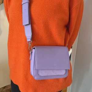 Ljuslila axelväska från C.W. - Säljer en stilren ljuslila axelväska från C.W. Väskan har ett brett axelband, lockstängning och en praktisk innerficka med dragkedja. Materialet är syntet med både släta och mockaliknande partier. Perfekt för att addera färg till din outfit!