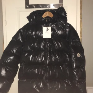 Svart dunjacka från Moncler (äkta) - Säljer en svart, glansig dunjacka från Moncler med alla lappar kvar och huva och dragkedja. Jackan har klassisk pufferstil och Moncler-logga på ärmen. Insidan har ett unikt serietidningsmönster och märkets signaturband vid nacken.