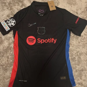 FC Barcelona svart bortatröja med Spotify-logga - Säljer en svart FC Barcelona bortatröja med röda och blå detaljer på sidorna, Spotify-logga på bröstet och Champions League-märke på ärmen. Tröjan har tryck med Lamine Yamal och nummer 19 på ryggen. Tillverkad i Dri-FIT material.