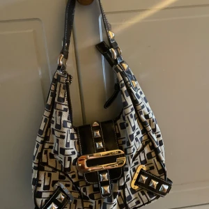 Mönstrad axelväska med nitar och spänne från Guess - Cool axelväska med svart och beige mönster, dekorerad med silvriga nitar och ett stort spänne framtill. Från guess!!