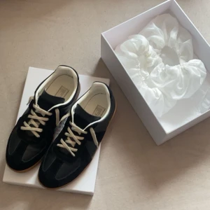 Svarta sneakers från Maison Margiela - Säljer ett par svarta sneakers från Maison Margiela med vita snören och beige sula. Skorna har en klassisk design med rund tå och paneler i både mocka och skinn. Perfekta för dig som gillar stilrena och tidlösa sneakers.