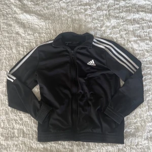 Svart Adidas track jacket med vita ränder - En fin adidas tracksuit jacka. Skicket är bra och har inga hål eller defekter.  Tyvärr är lapparna borta, men det är storlek 152 och passar 11/12 år.