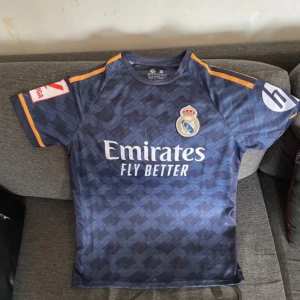 Real Madrid Bellingham 5 bortatröja - Mörkblå Real Madrid bortatröja med Bellingham 5 på ryggen. Tröjan har korta ärmar, subtilt mönster och gula detaljer på axlarna. Klubbmärke och sponsortryck på bröstet. Perfekt för fotbollsfans!