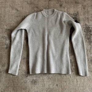 Ljusgrå ribbad tröja från Stone Island - Säljer en ljusgrå ribbad tröja från Stone Island med lång ärm och rund hals. Tröjan har en klassisk patch på ena ärmen och är tillverkad i ett stretchigt material som sitter snyggt på kroppen.