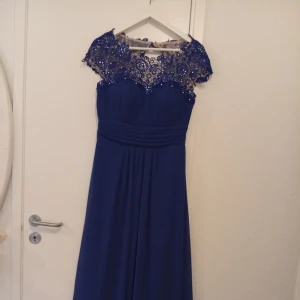 Blå långklänning med spetsdetaljer - Elegant blå långklänning med vacker spets upptill och transparent axelparti. Klänningen har markerad midja och faller snyggt ner mot golvet. Perfekt för bal eller festliga tillfällen.