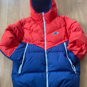 Nike röd och blå pufferjacka med huva - Säljer en färgstark pufferjacka från Nike i rött och blått. Jackan har huva, dragkedja framtill och två stora fickor med dragkedja. Perfekt för kalla dagar när du vill sticka ut lite extra.