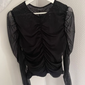 Svart mesh topp med rynkade detaljer - Säljer en svart topp med transparenta meshärmar och rynkade detaljer framtill. Toppen har långa ärmar i mesh som ger en cool och trendig look.