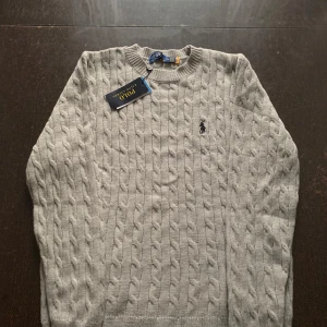 Grå kabelstickad tröja från Polo Ralph Lauren - Säljer en klassisk grå kabelstickad tröja från Polo Ralph Lauren. Tröjan har rund halsringning, lång ärm och den ikoniska broderade loggan på bröstet. Perfekt för en stilren och tidlös look.