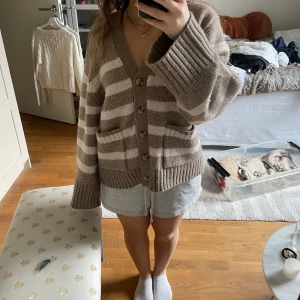 Beige och vit randig kofta från Lindex - Mysig kofta från Lindex i beige och vitt med breda ränder. Tröjan har v-ringning, knappar framtill och två fickor. Stickad i mjukt material och har långa ärmar samt ribbade muddar vid ärmslut och nederkant.