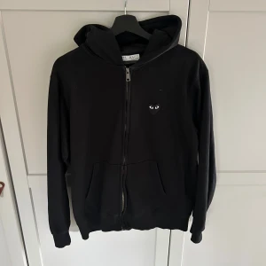 Svart hoodie från Comme des Garçons Play - Svart hoodie med dragkedja från Comme des Garçons Play. Ikoniskt hjärta med ögon på bröstet och stort hjärta med ögon på ryggen. Mjuk bomullskvalitet och två fickor framtill. Perfekt för en stilren och cool look.