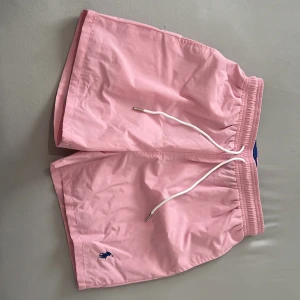Rosa badshorts från Polo Ralph Lauren - Helt unika badshorts som får allas ögon på dig i sommar. Snygga rosa badshorts från Polo Ralph Lauren med vit snörning i midjan och blå broderad logga på benet. Shortsen har elastisk midja och innerbyxa i mesh för extra komfort. Perfekta för strand eller pool! STOLEK S