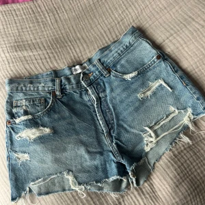 Blå jeansshorts från Zara - Säljer ett par blå jeansshorts från Zara med råa kanter och slitna detaljer. Klassisk femficksmodell med knapp och dragkedja framtill. Perfekta för sommaren och ger en cool, avslappnad look.